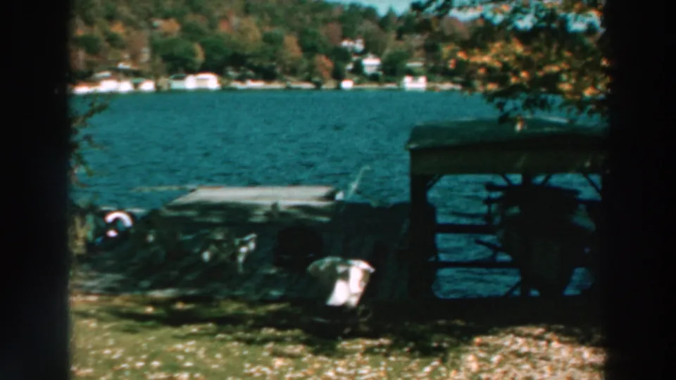 1964 Lake Winnebago frame showing a shaded shelter, dock, boats, grassy bank, trees, and distant shoreline homes.