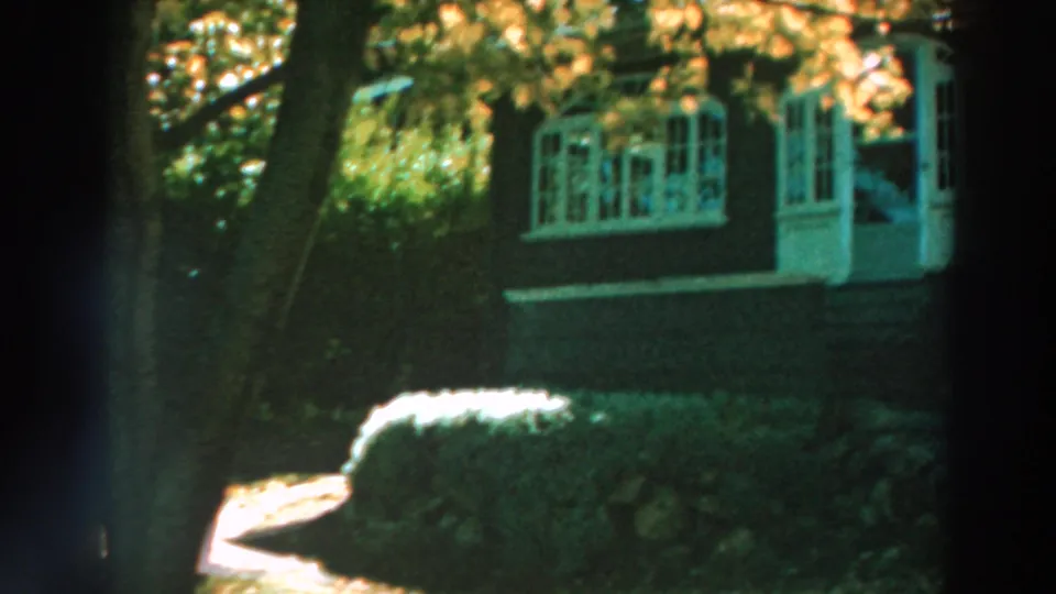 Lake Winnebago home-movie frame showing a red lakeside house with white-trimmed windows, trees, shrubs, and warm sunlight.