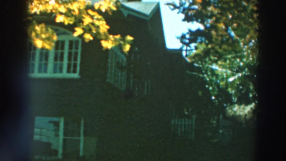 1964 Lake Winnebago home-movie frame showing the side of a dark house, a white window, roofline, trees, and shaded yard vegetation.