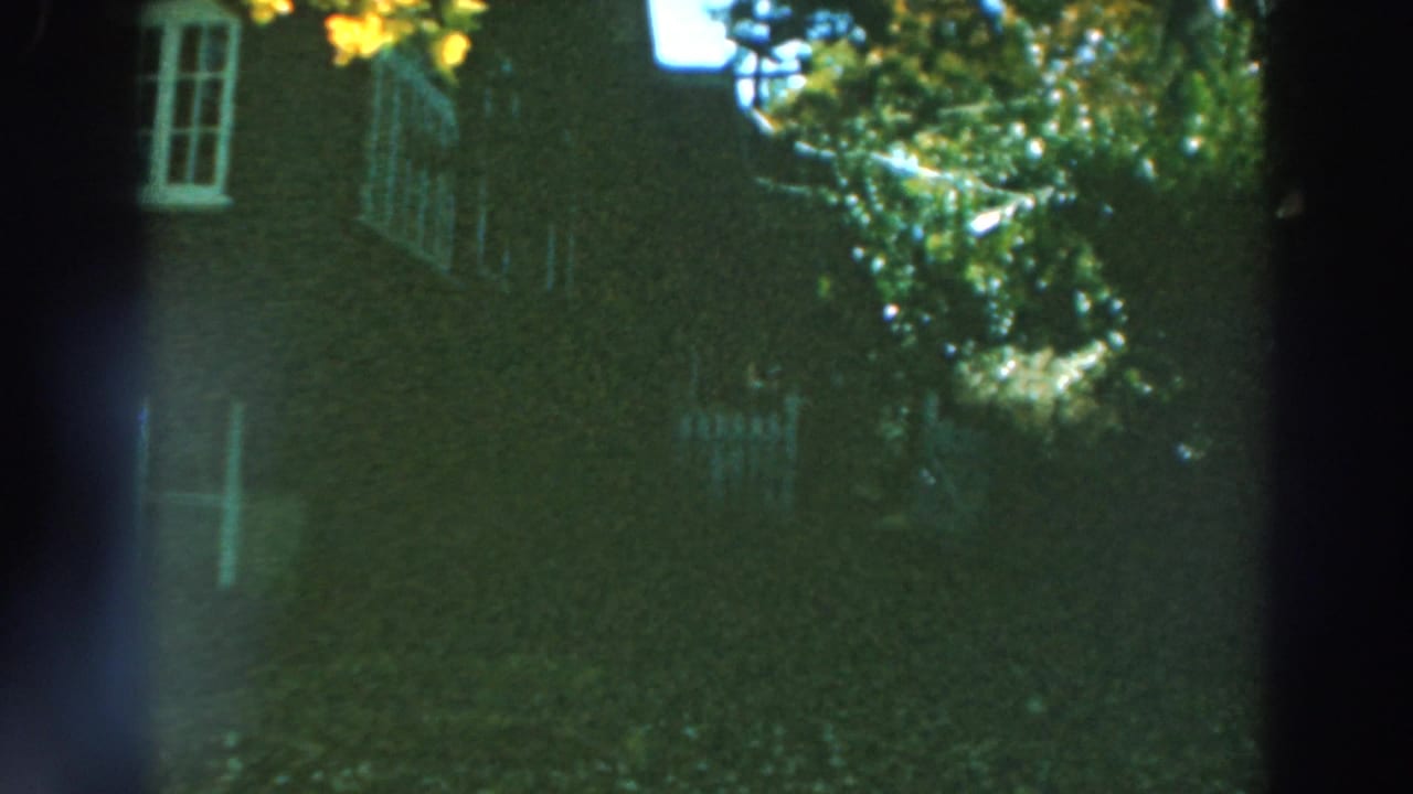 1964 Lake Winnebago home-movie frame showing a shaded brick house, white window trim, trees, and a white fence.