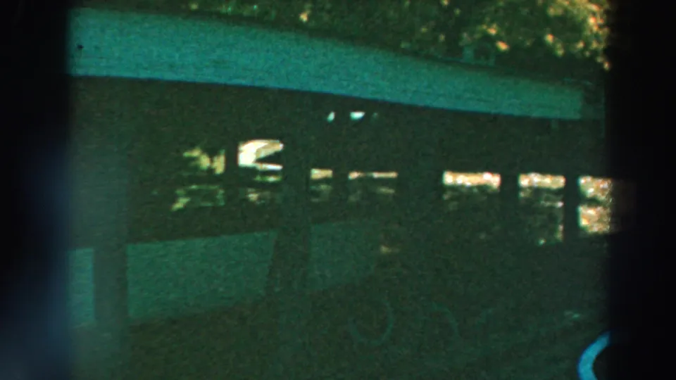 1964 Lake Winnebago 8mm frame showing a white boat stored beneath a covered wooden structure, surrounded by trees and strong shade.