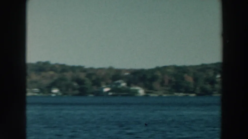 1964 Lake Winnebago boat view with distant shoreline buildings, wooded slopes, blue water, and pale sky.