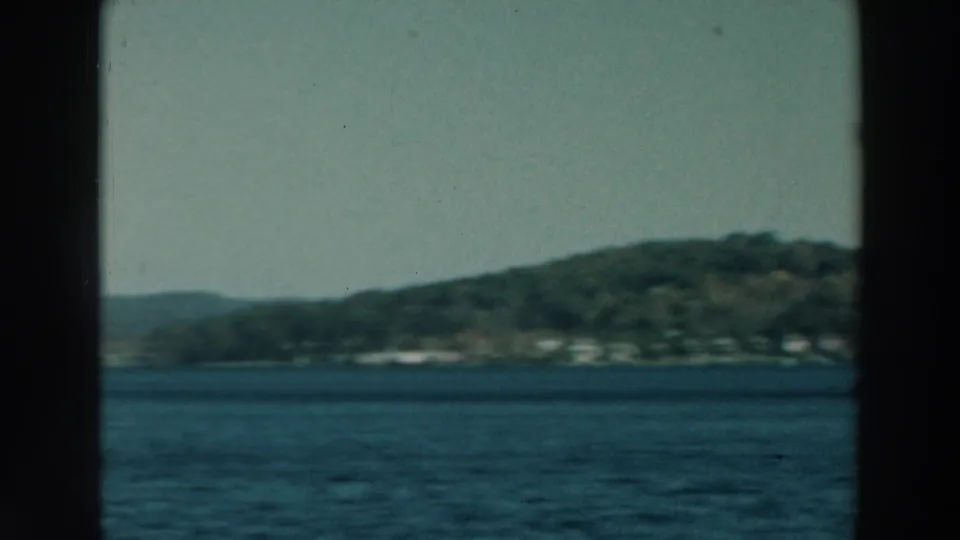 1964 Lake Winnebago view with blue water, forested hills, distant shore buildings, and dark window mullions in the foreground.