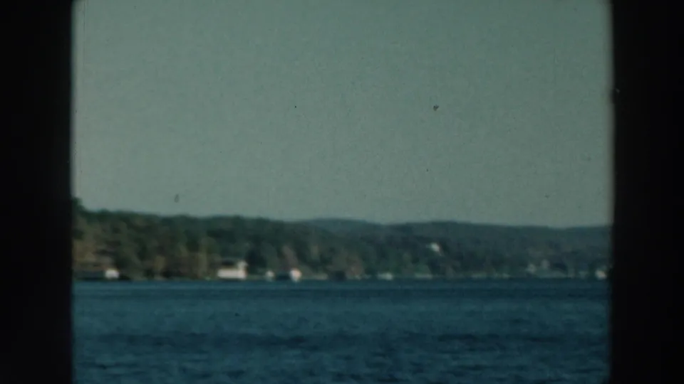 1964 Wisconsin lake scene showing water, low wooded hills, scattered shoreline structures, and a dark center window divider.