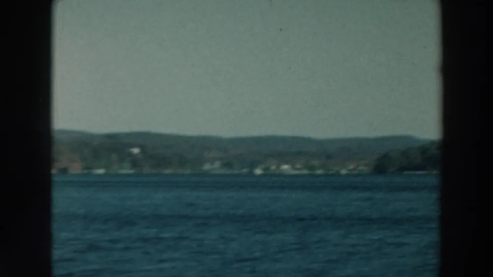 1964 Lake Winnebago footage looking across blue water toward a low, wooded shoreline partly obscured by window framing.