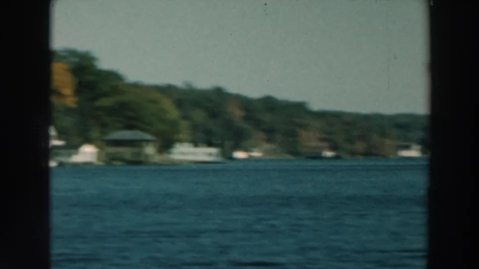 1964 Wisconsin lake view with wooded shoreline, scattered small buildings, blue water, and dark window framing at the edge.
