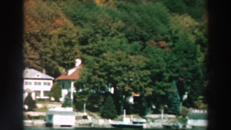 1964 Lake Winnebago home-movie frame showing white shoreline houses, docks, boats, and dense trees across the water.