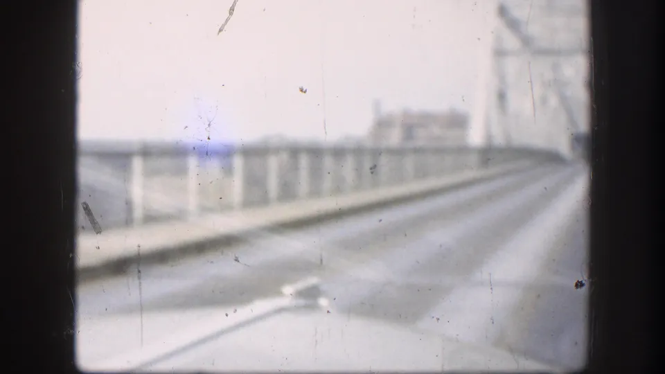 From a moving vehicle on a bridge, the camera looks ahead along the roadway with railings and blurred bridge structure visible.