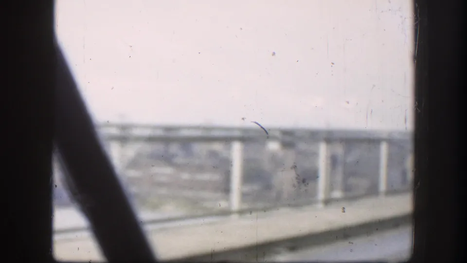 A dark vehicle-window post and diagonal interior edge frame the bridge railings and concrete supports during the river crossing.