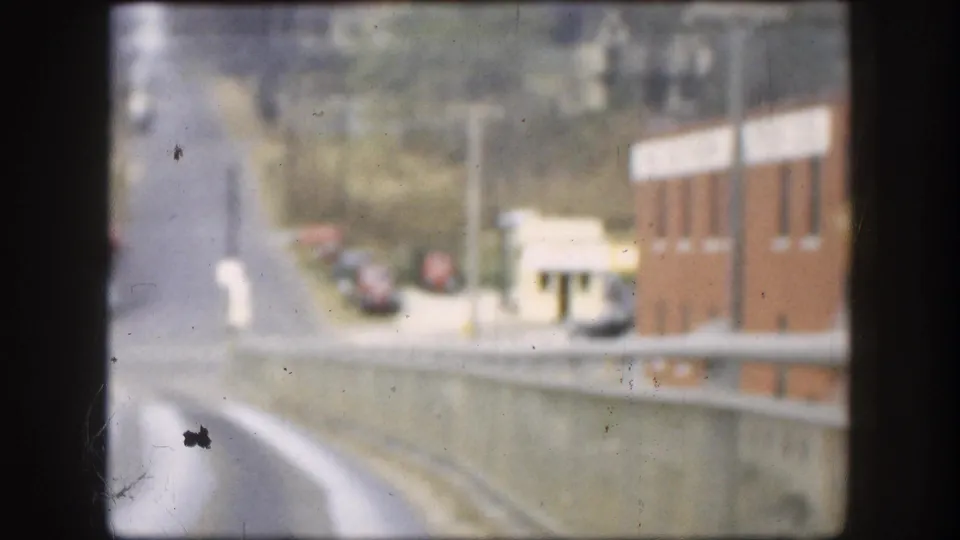 From inside the car, the road drops away from the bridge near a red brick building, parked cars, and roadside poles in Kansas.