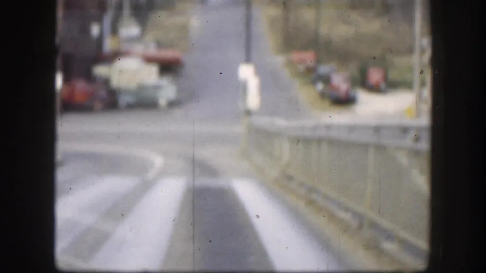 A vehicle-window view shows the bridge exit, road markings, guardrails, parked cars, and low buildings in a 1958 Kansas street scene.