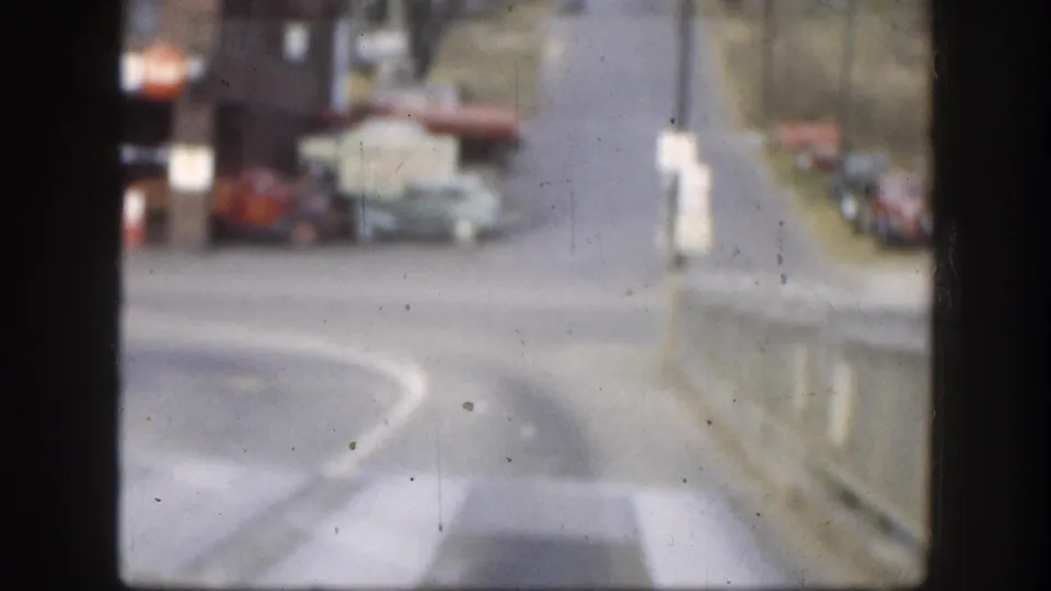 Kansas 1958 8mm frame showing a blurred street scene with parked mid-century cars, a road curve, and dark window-like borders.