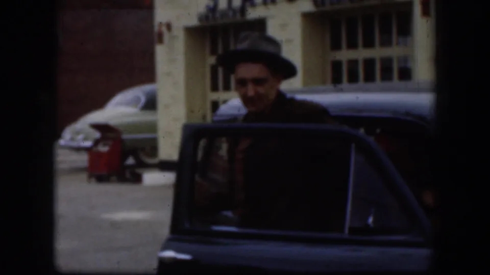 Adult man in a dark hat stands by an open sedan door in front of a building with parked mid-century cars.