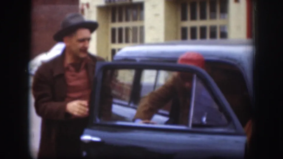 Two adult men gather by a dark sedan, with one man in a hat outside and another partly visible at the open door.