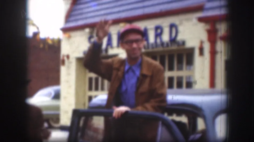 Man in a red cap and glasses waves beside a dark sedan, with a Standard sign visible on the building behind him.