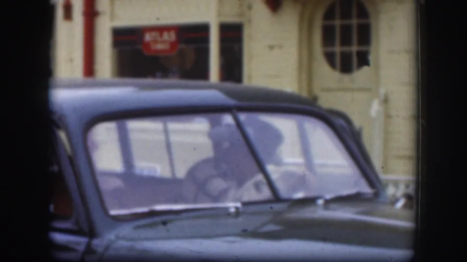 A dark mid-century car is seen close up in Kansas, with occupants visible through the windows and a service-station building behind it.