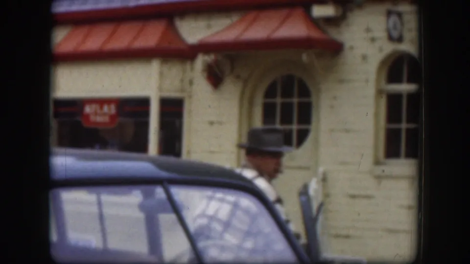 A man in a brimmed hat moves beside the dark car in the 1958 Kansas street scene, with the cream facade and red signage behind him.