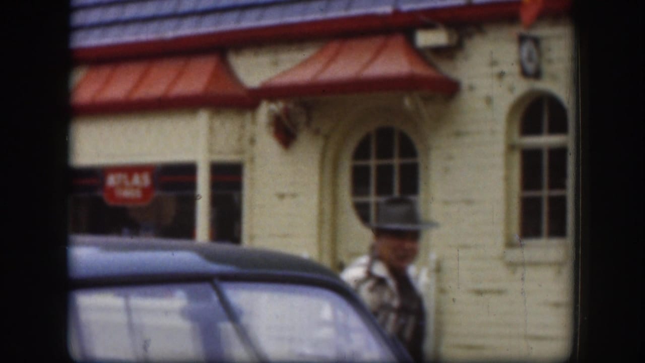 1958 Kansas home-movie frame showing a man near a vintage car in front of a cream building with red awnings and an ATLAS sign.