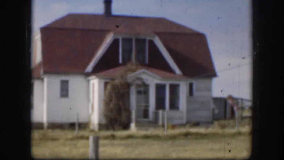 Late-1950s Kansas home-movie frame of the Bjerke home exterior, with a red roof, dormer windows, porch entry, and open land around it.