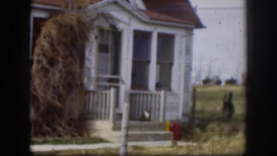 1958 Kansas frame focused on the house porch, white railings, dried brush, and nearby yard details.