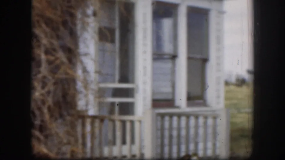 Close 1958 Kansas 8mm view of a screened porch with white railings and dried brush leaning beside the house.