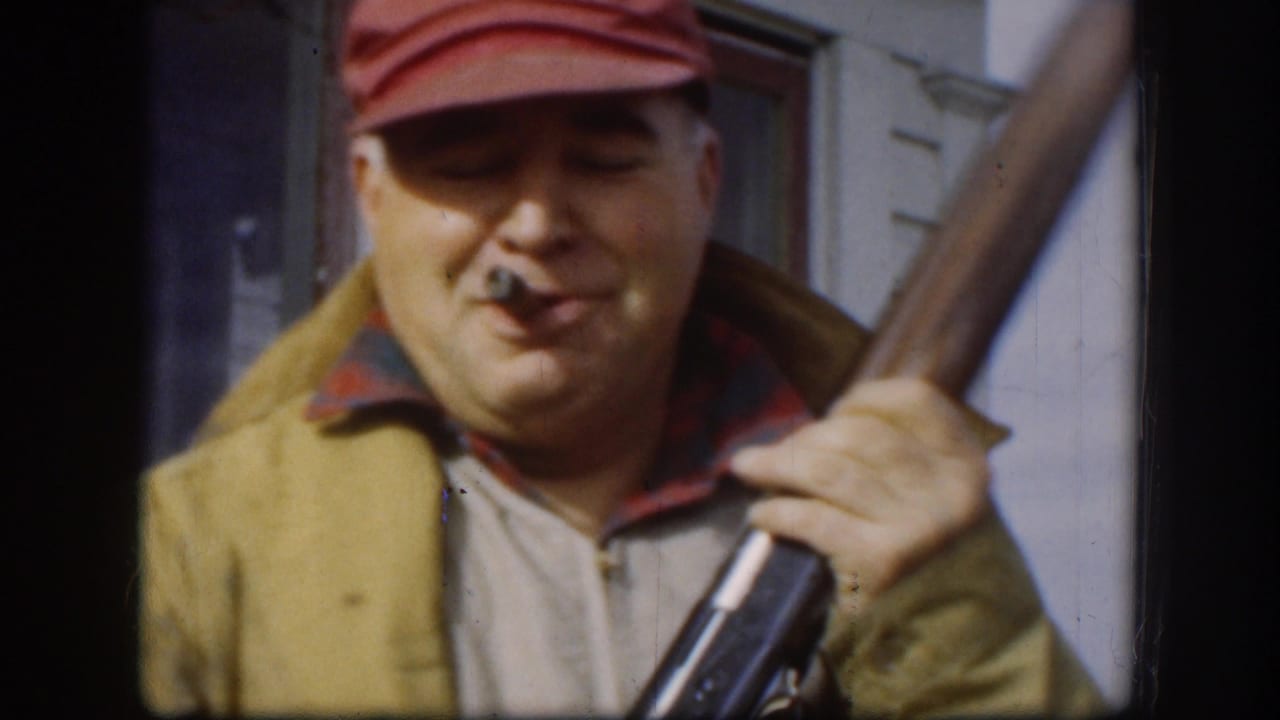 Adult man in a red cap smokes a cigar and holds a shotgun beside a building in 1958 Kansas home-movie footage.