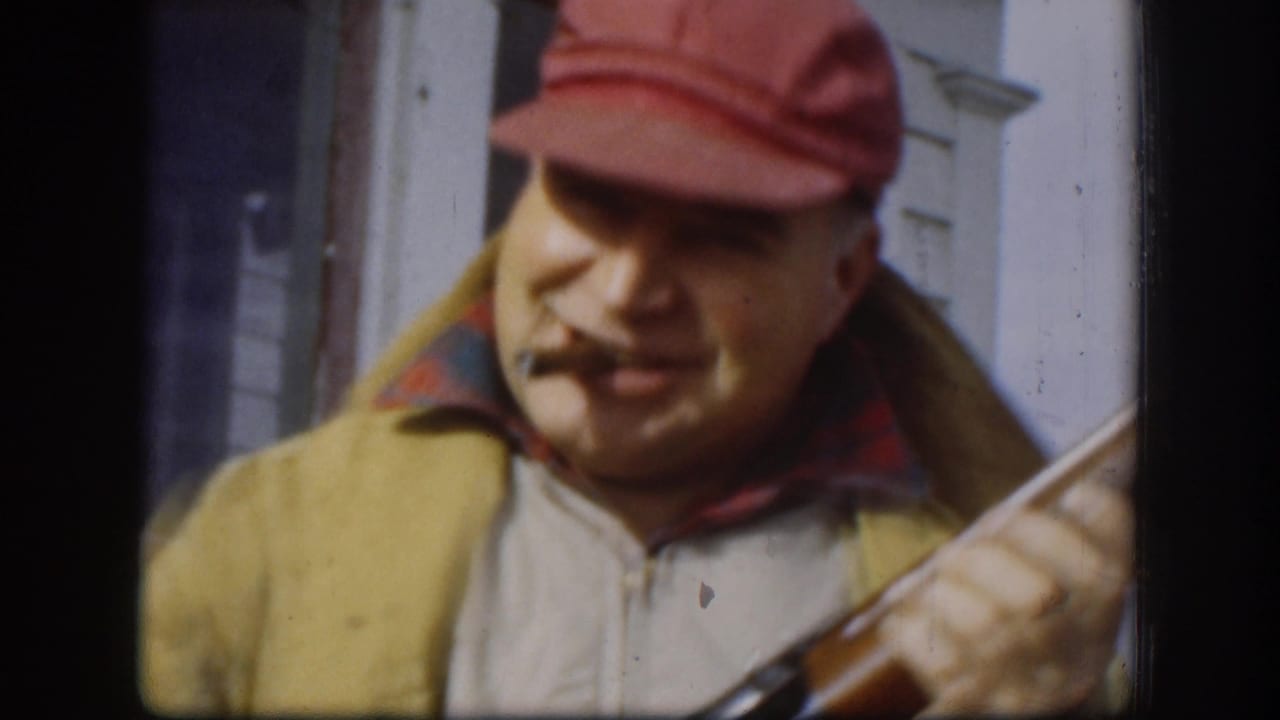 Man in red cap with cigarette holds a shotgun near white siding.