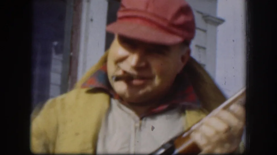 In 1958 Kansas home-movie footage, a man in a red cap and tan vest smiles with a cigarette while holding a shotgun near a white building.