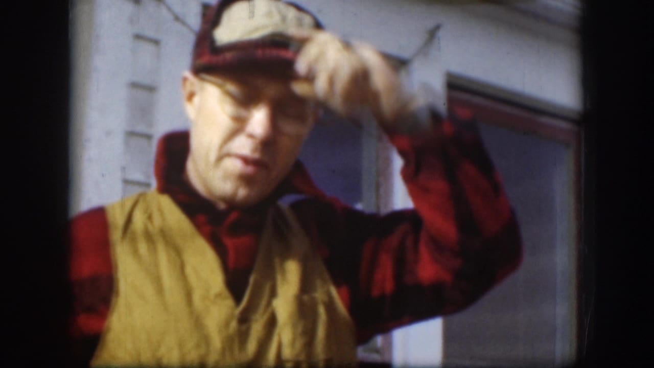 In a 1958 Kansas home-movie frame, an adult man in glasses, plaid shirt, tan vest, and cap raises his hand near the brim.