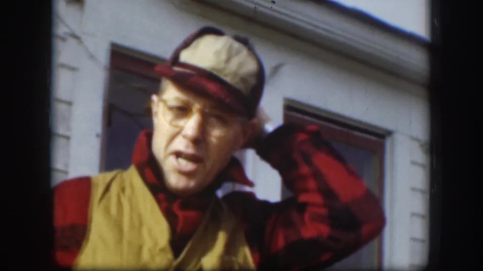A candid 1958 Kansas frame shows the man tilting his cap, his glasses and plaid shirt visible beside a dark window frame.