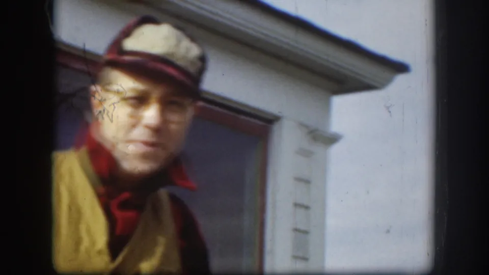 The man moves partly out of frame in the 1958 Kansas home movie, with his cap, glasses, plaid shirt, and tan vest still visible.