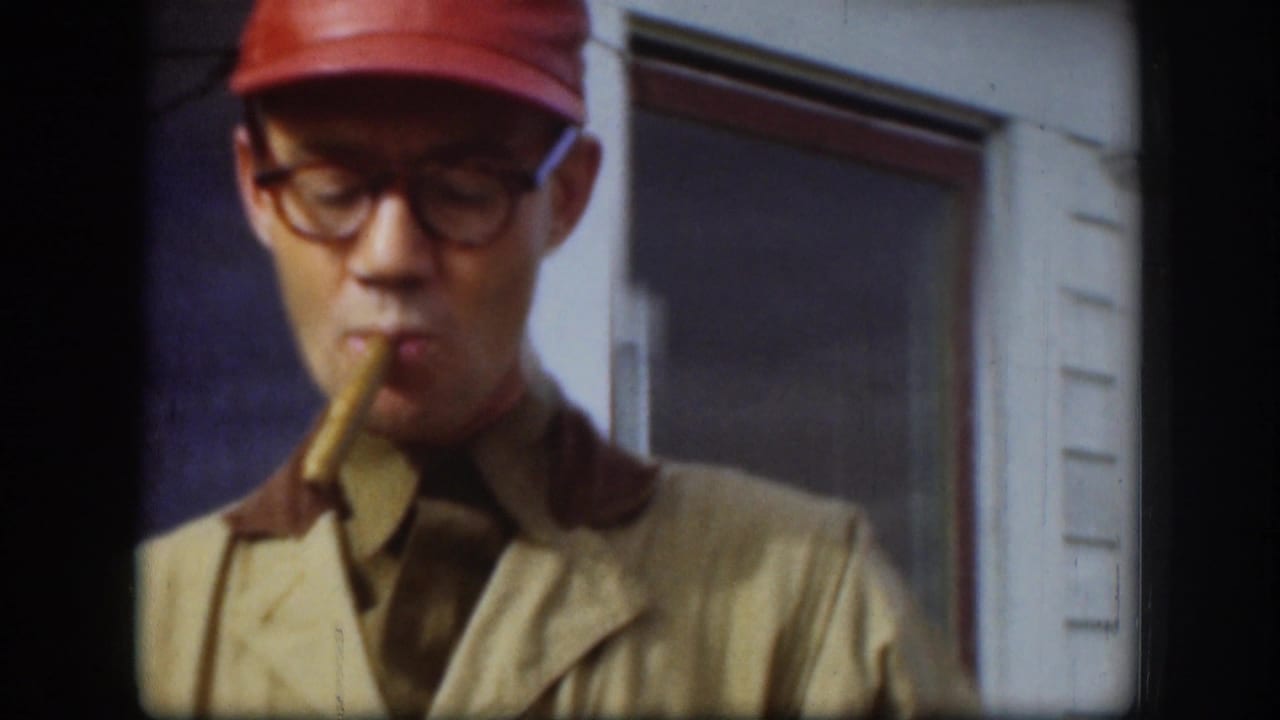 Man in a red cap and glasses holds a cigar outside a white building.