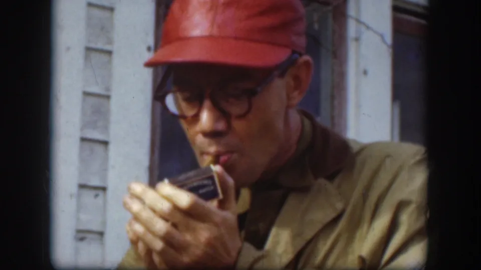 1958 Kansas 8mm frame showing the man in a red cap and glasses focused on lighting his cigar beside a white building.