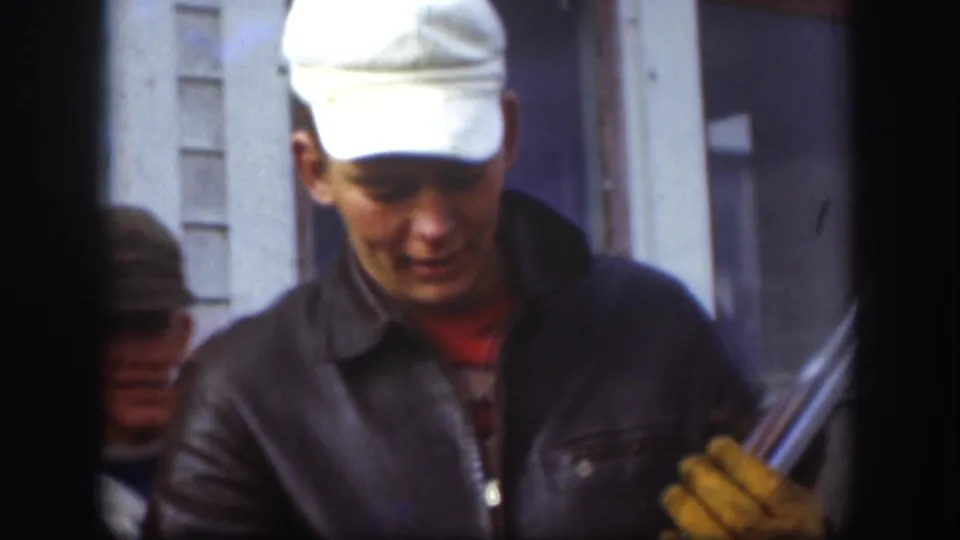 1958 Kansas 8mm frame showing a man in a white cap and dark jacket holding a long gun near a white building.