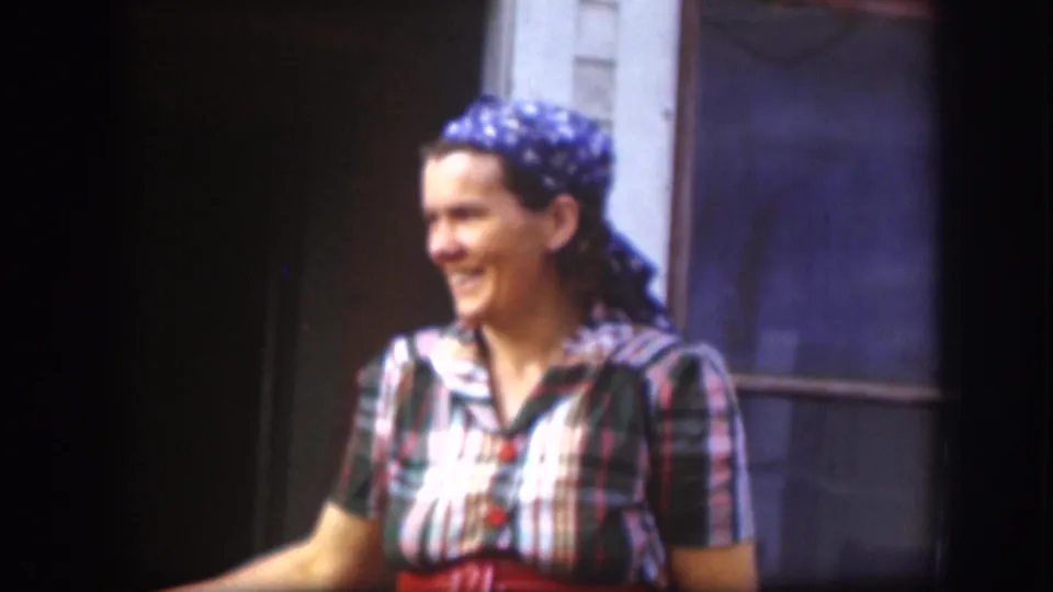 Adult woman in a plaid dress and blue headscarf smiles outdoors in 1958 Kansas home-movie footage.