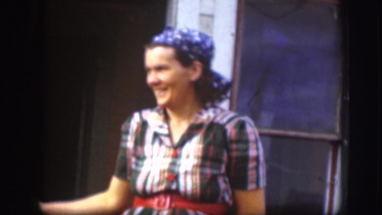 In 1958 Kansas home-movie footage, a woman in plaid clothing and a blue headscarf smiles beside a doorway.