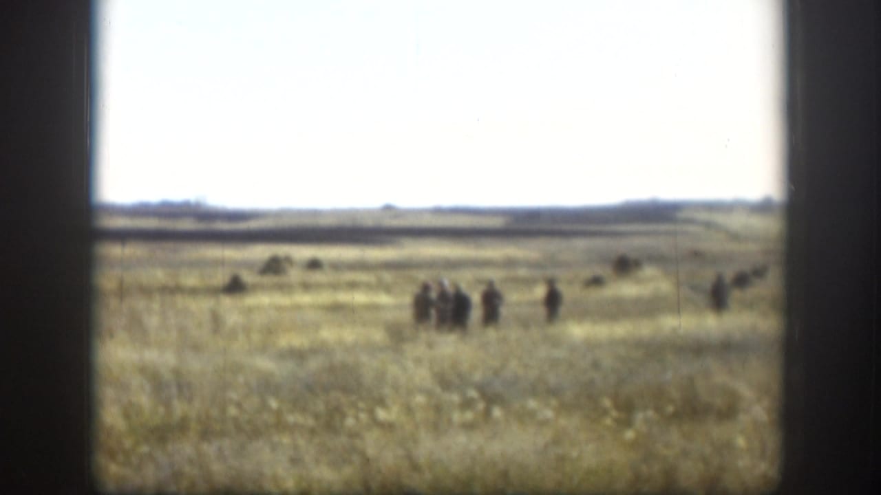 A wide Kansas field view shows small distant figures gathered across the grass beneath a pale sky.