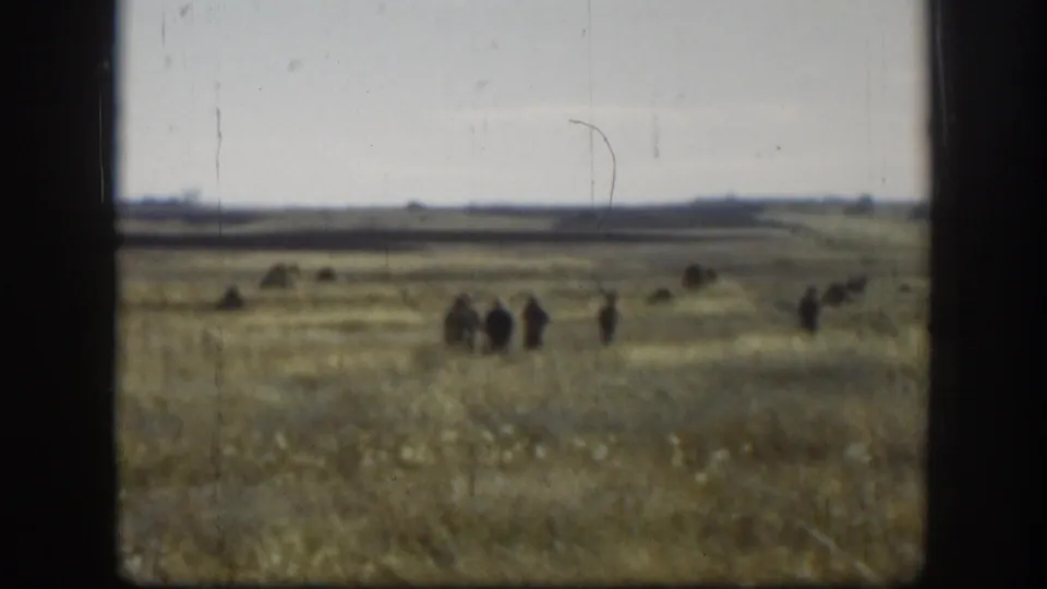 Several distant figures appear spread across an open Kansas field in soft 8mm color footage.