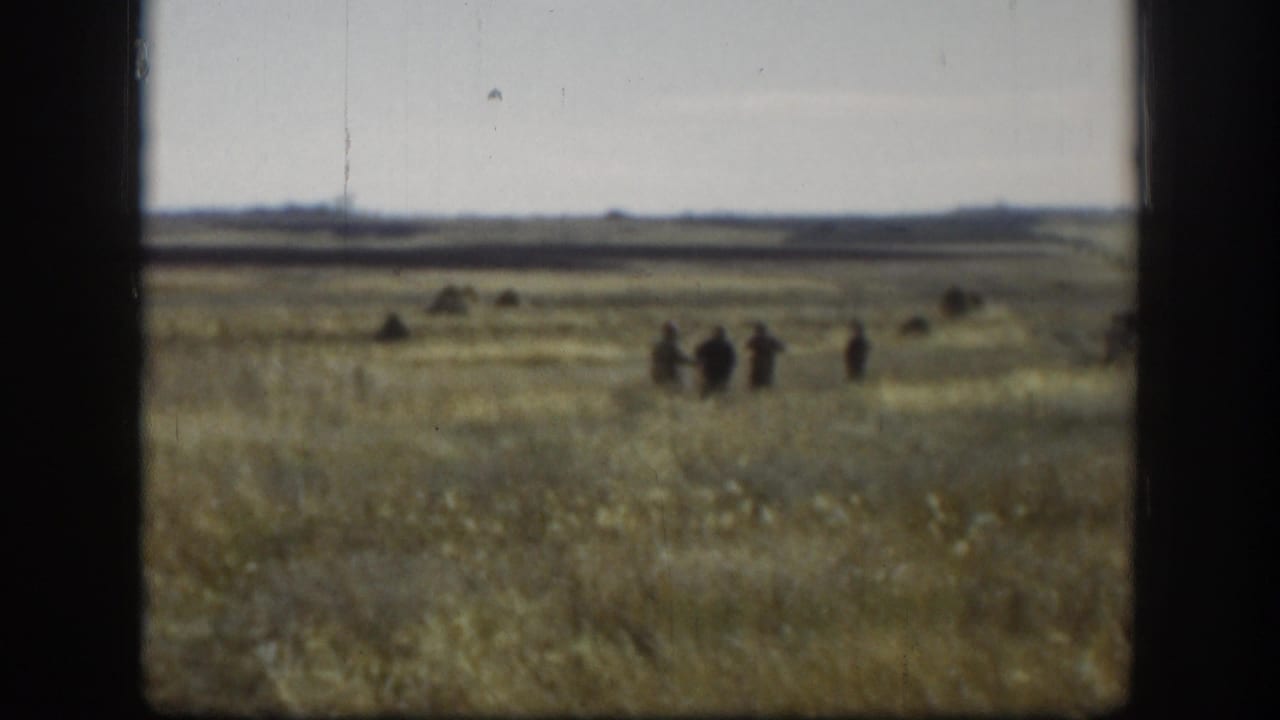 Distant hunters standing in a wide grassy field under a pale sky.