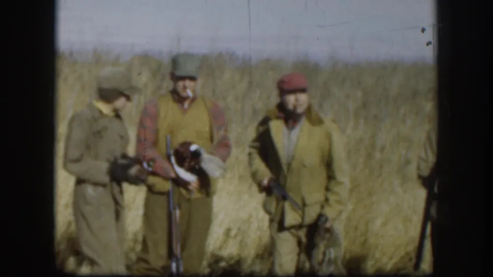 In 1958 Kansas footage, hunters in caps and vests stand in tall grass while one man holds a harvested duck near a shotgun.