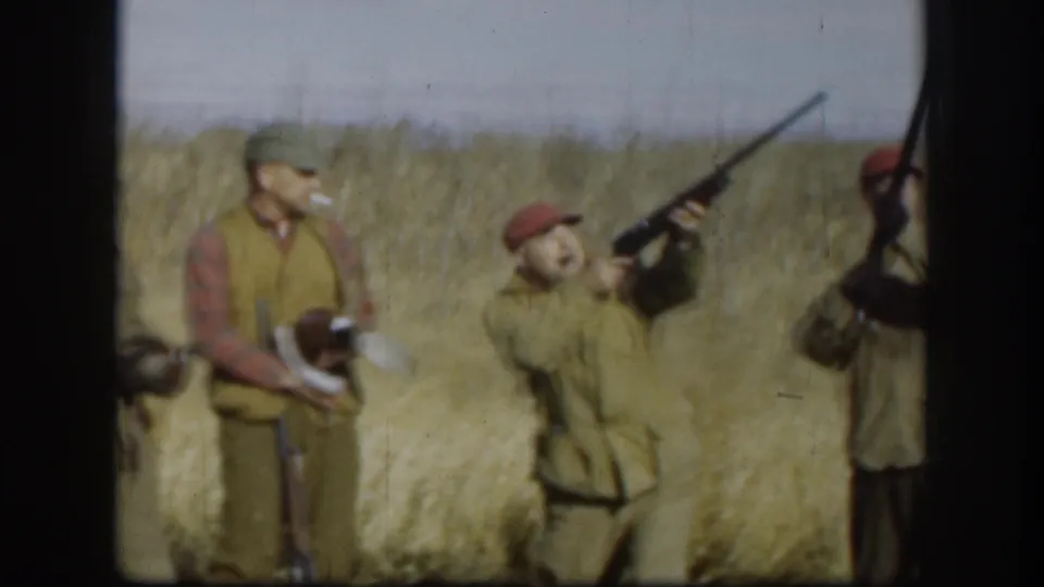 Adult hunters in a dry Kansas field hold shotguns and ducks, with one man tilting his head upward as another gun is raised nearby.