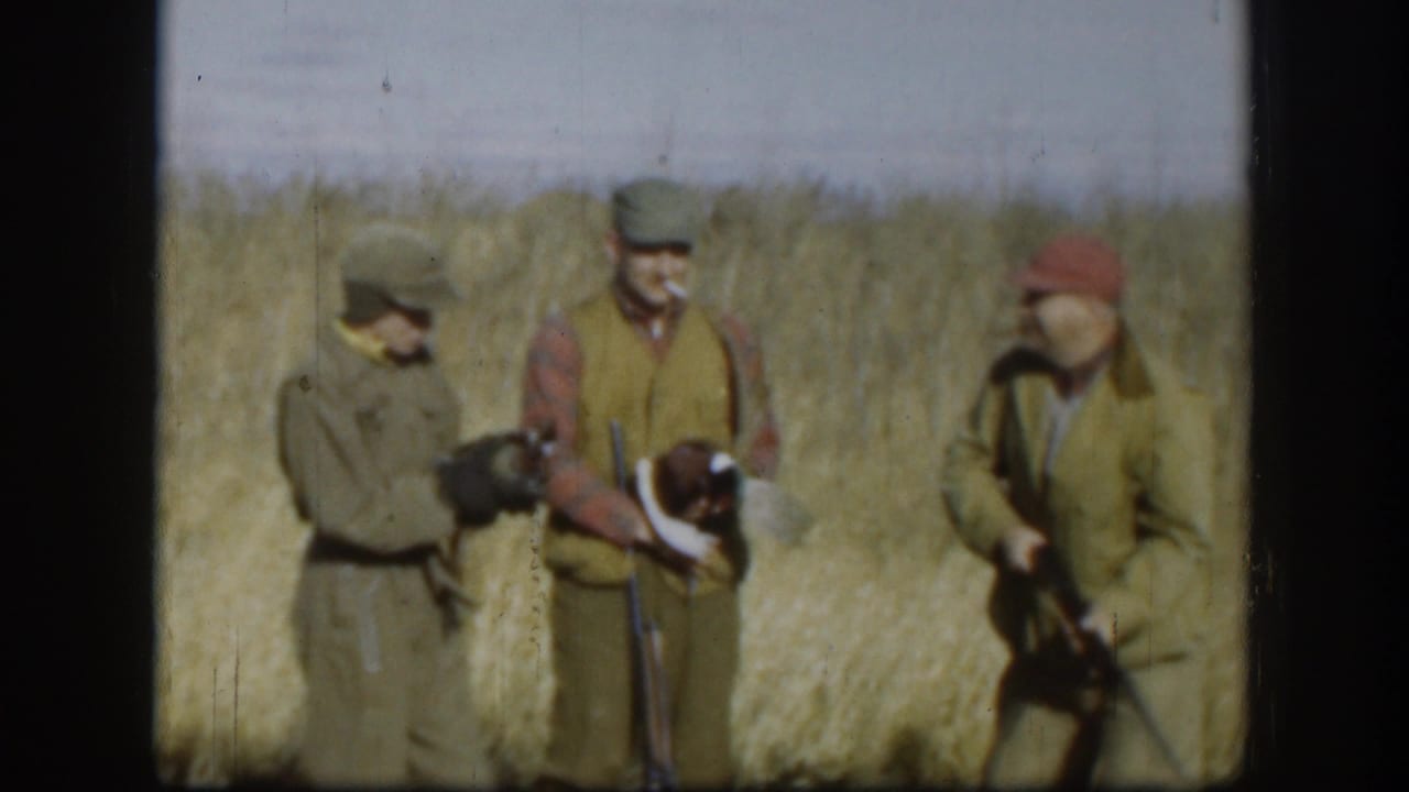 Three men in hunting clothes stand in a dry field, with rifles and a possible game bird visible.