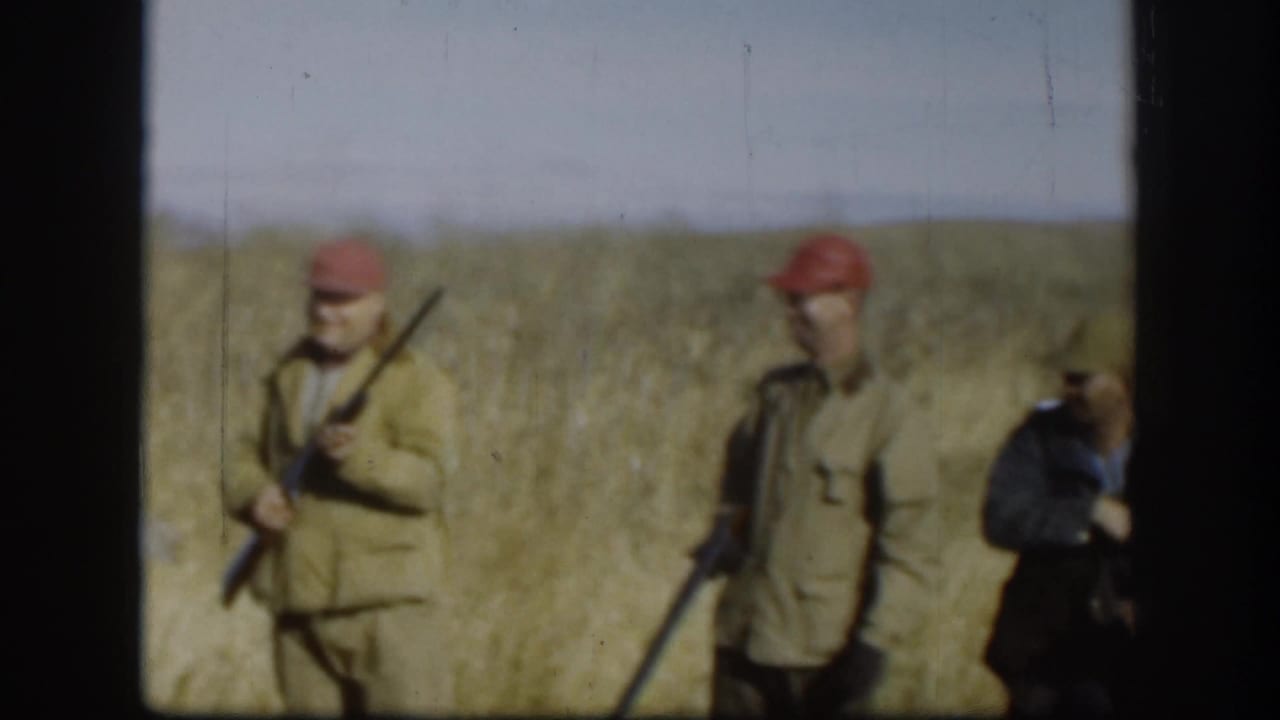 Two adult hunters in caps and tan jackets stand with rifles in a Kansas field, with another figure partly cropped at the right edge.