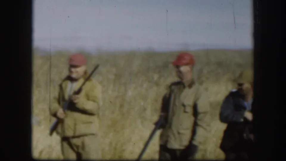 Two adult hunters in caps and tan jackets stand with rifles in a Kansas field, with another figure partly cropped at the right edge.