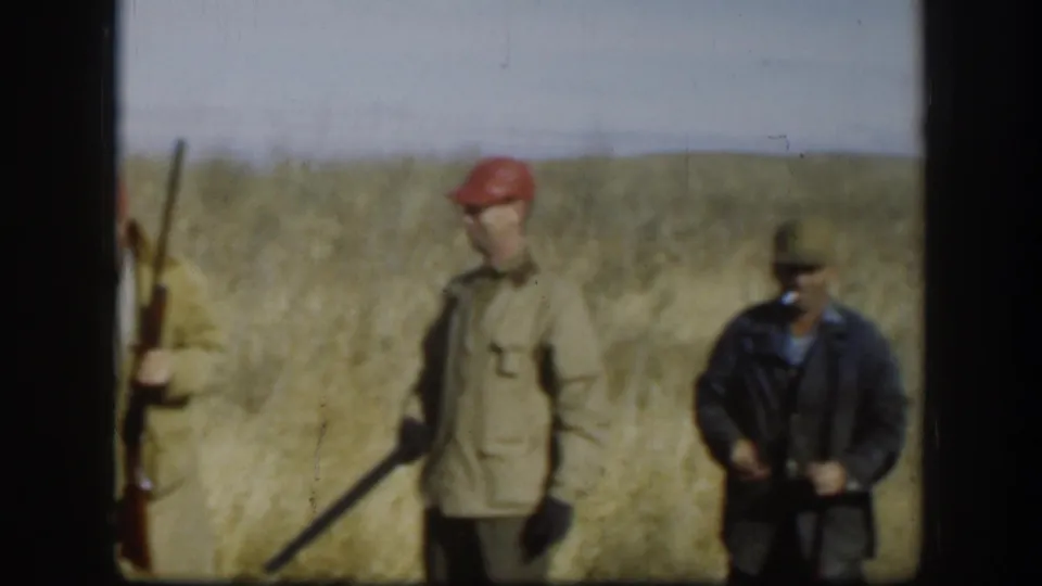 A hunter in a red cap and tan jacket holds a rifle in a dry Kansas field while another adult man stands nearby in darker clothing.