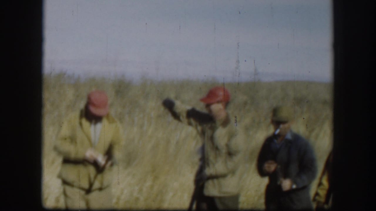 Adult hunters in caps stand in a dry field, with shotguns and one man smoking.