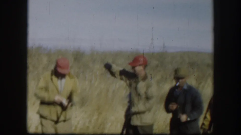 Kansas 1958 home-movie frame showing adult hunters in caps and jackets standing in tall dry grass with shotguns, one appearing to smoke.