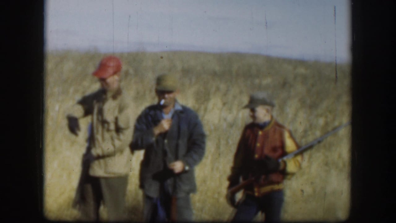 Kansas 1958 frame of three adult hunters in caps and jackets standing in tall grass, with shotguns visible and one man smoking.