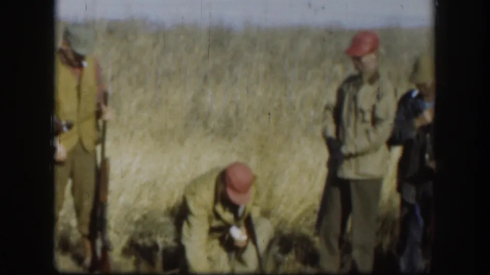 Kansas 1958 frame with hunters in caps and jackets standing in a dry field, one bending down while shotguns remain visible.