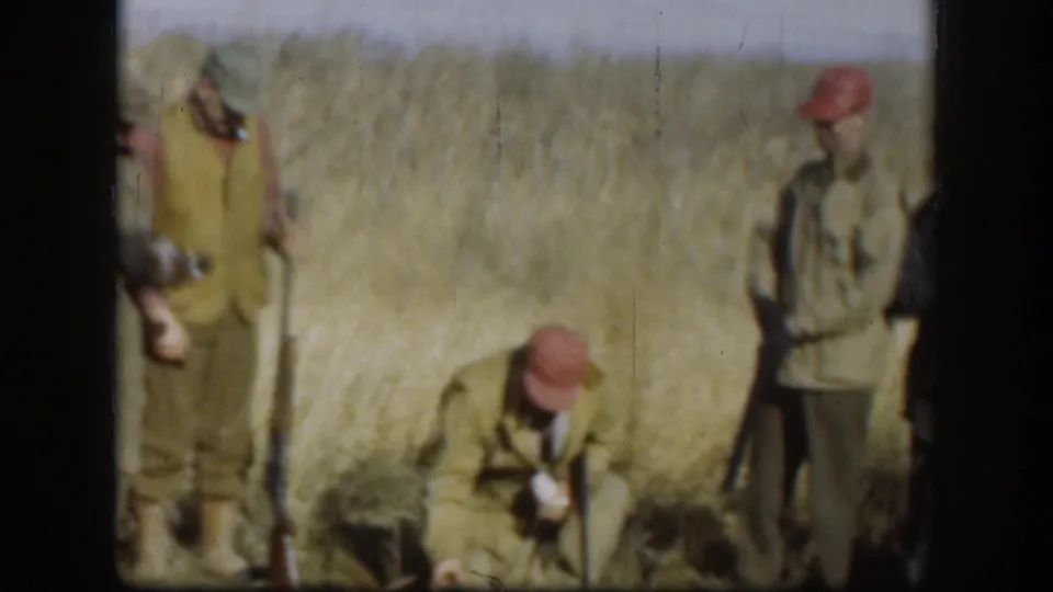 1958 Kansas home-movie frame showing men in a rural field, with one kneeling and others standing in tall grass.
